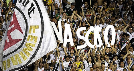 Torcida organizada do Vasco com bandeira, apoiando e mostrando entusiasmo no estádio, com muitos torcedores vestindo as cores do clube.