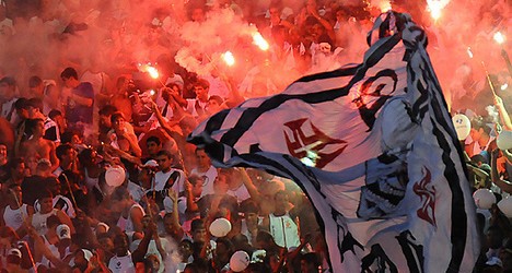 Vascaínos comemoram com bandeira e sinalizadores em estádio cheio. Atmosfera de entusiasmo e paixão pelo clube Vasco da Gama, durante partida ou evento especial.