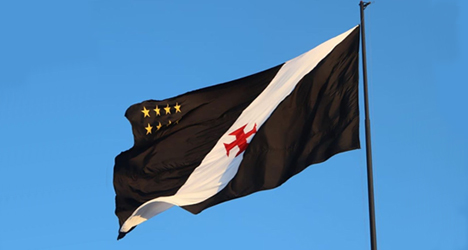 Bandeira do Vasco da Gama ao vento contra o céu azul claro, destacando as cores preta, branca, vermelha e as estrelas douradas, símbolo do clube carioca.