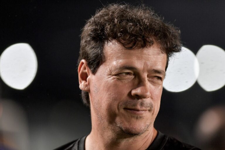 Homem sorrindo com cabelo castanho escuro e fundo de luzes brancas.