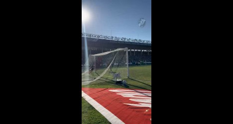 Futebol em campo com arquibancada ao fundo.