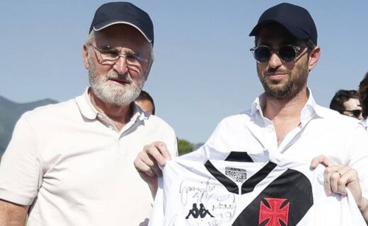Homem segurando camisa do Vasco com assinatura, ao lado de outro homem com óculos e camisa branca.