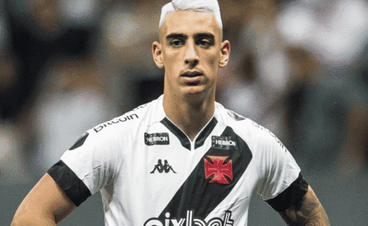 Imagem de jogador do Vasco com camisa branca e faixa preta, com expressão séria, em campo de futebol.