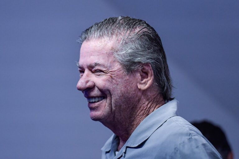 Aposentado sorrindo, cabelo grisalho, fundo azul.