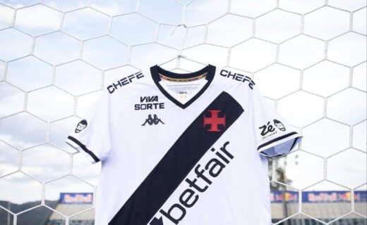 Estandarte de uma camisa do Vasco da Gama pendurada em uma rede de futebol, em um estádio de futebol ao ar livre, com grama verde e céu azul ao fundo.