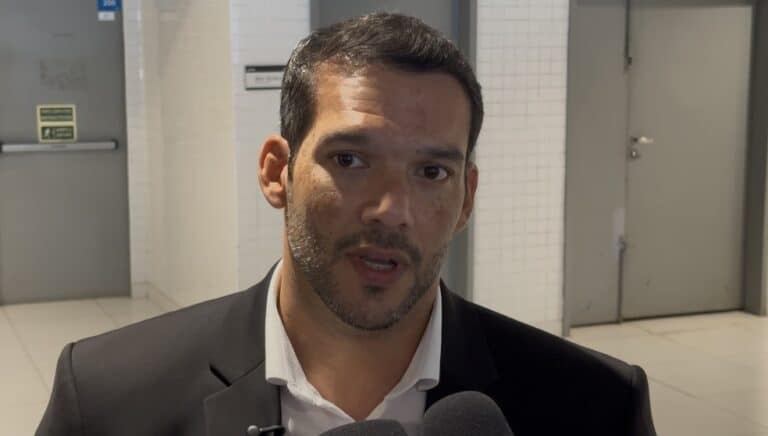 Vestido de terno preto, falando em entrevista, fundo de clínica ou consultório.