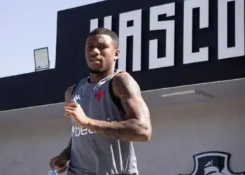 Novo jogador do Vasco, com uniforme cinza, em frente ao painel do clube.