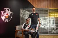 Imagem de dois homens na sala de eventos do Vasco, um sentado e outro em pé ao lado, fundo com logo do clube e mesa com computador e cadeiras | Jogador e treinador do Vasco na sala de eventos do clube, celebrando uma conquista, com o logotipo do time ao fundo e ambiente de trabalho | A imagem mostra um jogador e um treinador do Vasco em ambiente interno, possivelmente em uma cerimônia ou reunião oficial, com o logotipo do clube visível no fundo. O jogador está sentado em uma cadeira e o treinador ao lado, ambos sorrindo. O cenário inclui uma mesa com computador e cadeiras, destacando o ambiente institucional do clube | Relevância: relevante para conteúdo de notícias ou materiais institucionais do Vasco, reforçando a presença e atividades do clube em eventos internos.