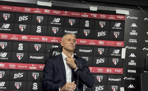 Imagem de um homem em pé, usando terno, sentado na mesa de uma coletiva de imprensa. fundo com logos do São Paulo Futebol Clube, patrocinadores e marcas associadas ao time.