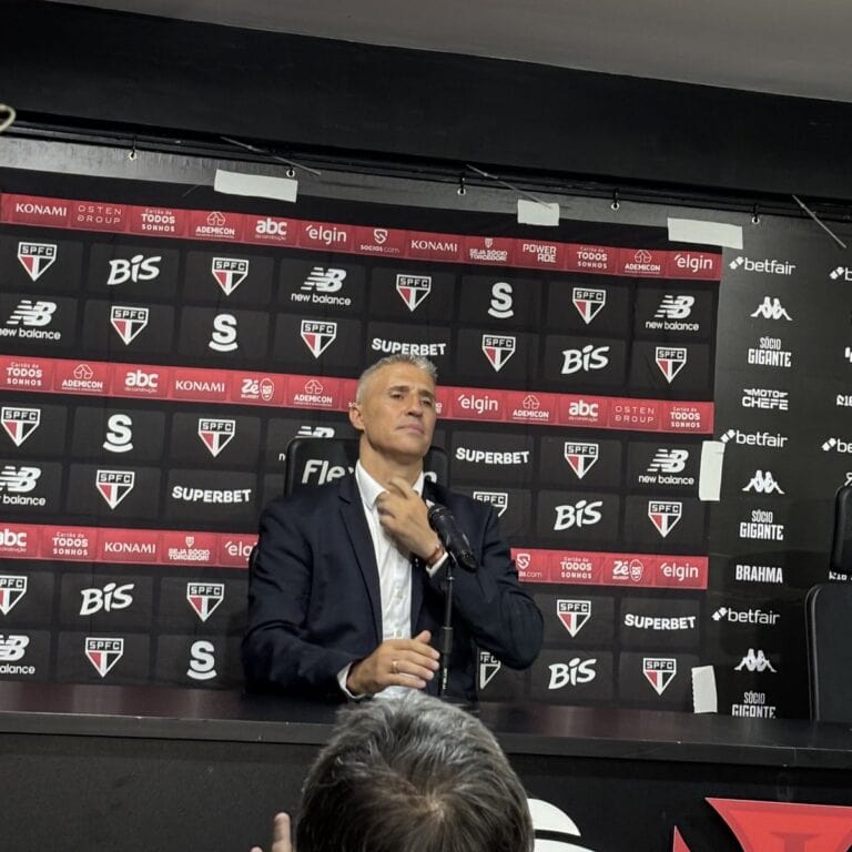Imagem de um homem em pé, usando terno, sentado na mesa de uma coletiva de imprensa. fundo com logos do São Paulo Futebol Clube, patrocinadores e marcas associadas ao time.