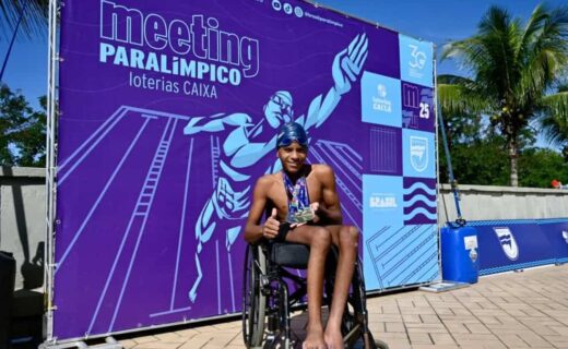 Atleta em cadeira de rodas com medalhas, sorrindo, em fundo de evento esportivo.