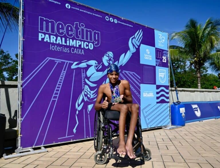 Atleta em cadeira de rodas com medalhas, sorrindo, em fundo de evento esportivo.