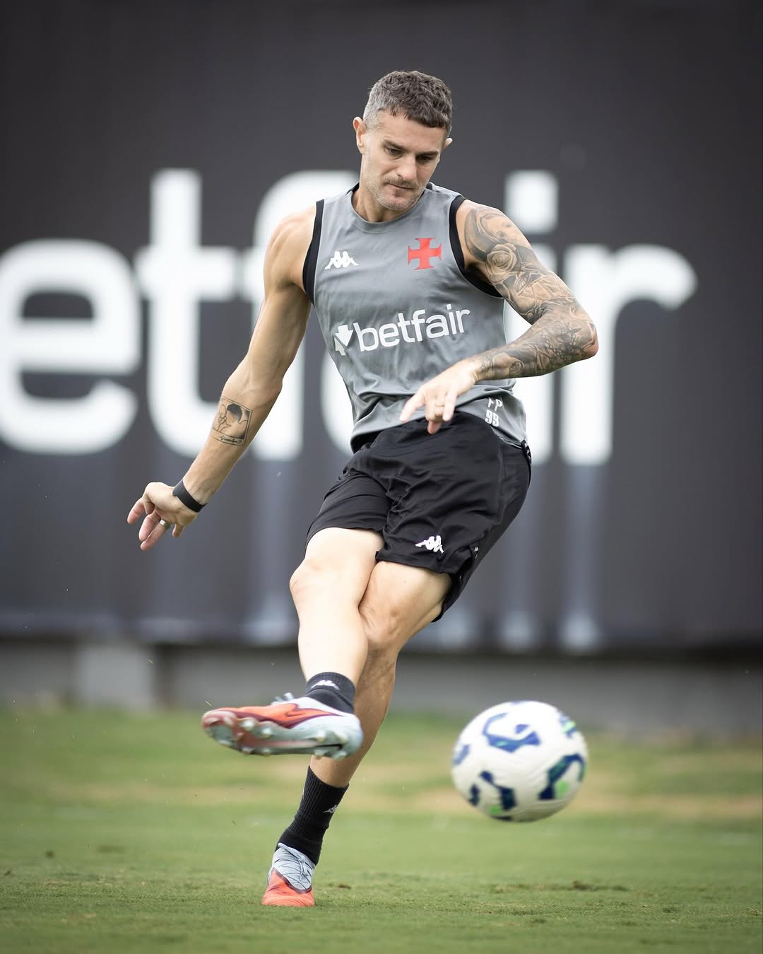 Jogador de futebol treinando com a bola no campo, vestindo uniforme cinza e preto.