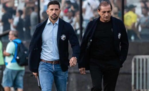 Treinadores do clube Vasco, usando jaquetas e mochila, caminhando pelo gramado enquanto os torcedores aparecem ao fundo no estádio.