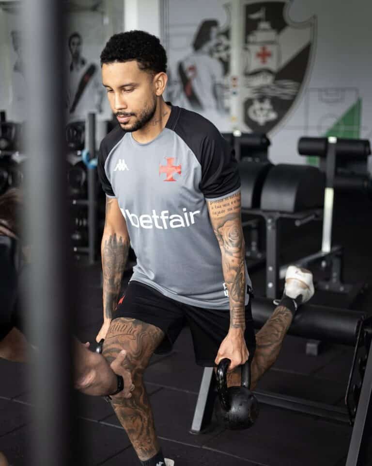 Este homem com barba e tatuagens treina no ginásio vestindo camisa cinza e preta do Vasco, segurando halteres durante exercício focado.