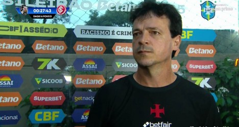 Treinador do Vasco durante entrevista após treino, com fundo de patrocinadores e ambiente esportivo ao redor.