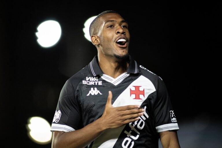 Jogador do Vasco comemorando gol vestindo uniforme preto com detalhes brancos, símbolo do clube na camiseta, fundo escuro com luzes difusas.