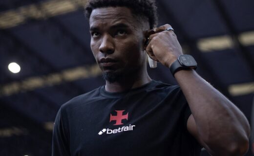 Homem negro com camiseta do Vasco, ajustando fone de ouvido no estádio.