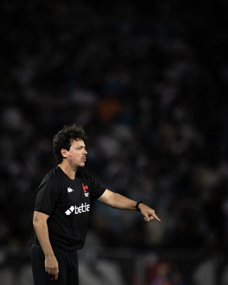 Treinador do Vasco da Gama em ação durante jogo, vestindo uniforme preto com logo de patrocinador, com expressão concentrada em estádio lotado.