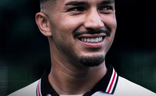 Texto alt: Fotografia de um homem sorridente com cabelo escuro, barba por fazer, usando camisa de time de futebol, com fundo desfocado escuro e verde.