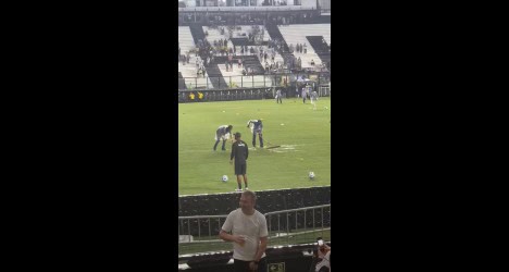 Jogadores do Vasco treinando na academia, com foco no campo de futebol cheio de marcas de treino. Torcedores podem ver a preparação do time.