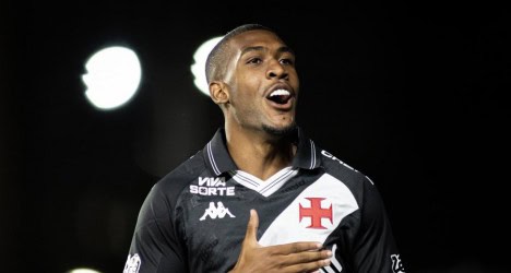 Atacante do Vasco comemorando gol com a camisa do time, com fundo escuro e iluminação focada, expressando alegria e entusiasmo na partida.