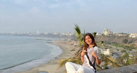 Vascaína sorridente com uniforme do Vasco de frente para o mar na praia ao entardecer, cenário urbano ao fundo.