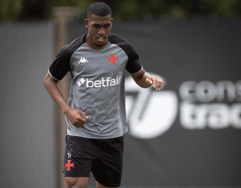 Texto alternativo: Jogador do Vasco treinando, vestindo uniforme cinza e preto com o escudo do time, focado na atividade, em ambiente de campo de futebol.